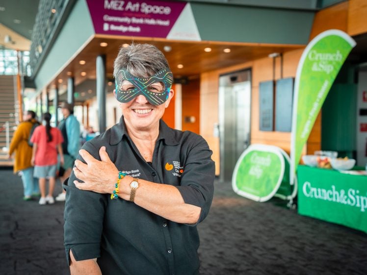 Image of A smiling woman wearing a colourful mask and black shirt stands indoors, posing with one arm crossed over her chest. Behind her are people, a table with drinks, and green banners that read Crunch&Sip.