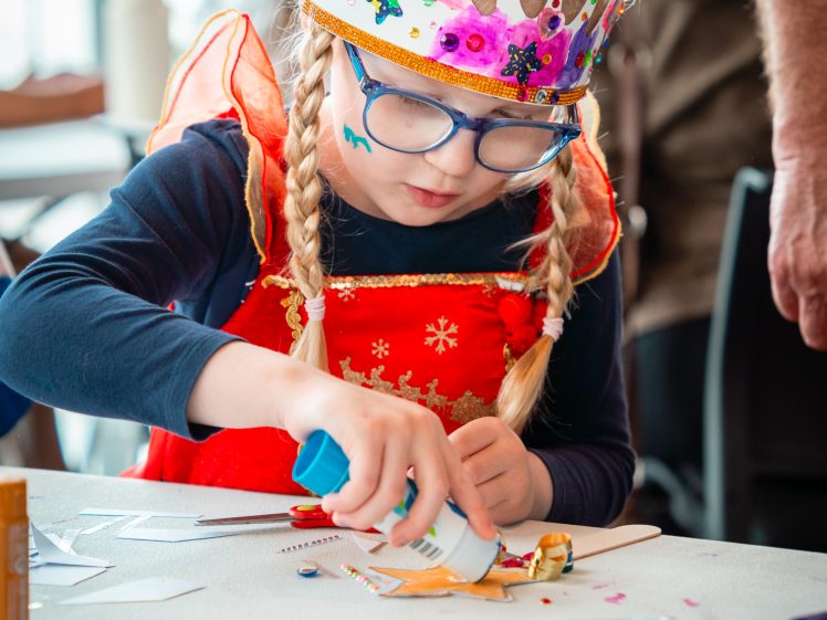 Image of A young child with fair plaits and glasses wears a decorated paper crown and red costume, concentrating whilst gluing papers and crafting at a table covered with art supplies.