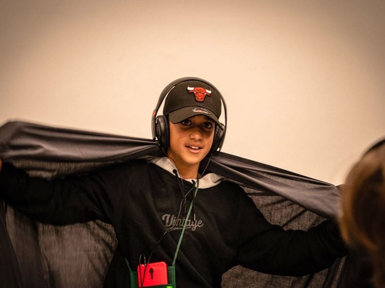 Image of A boy wearing headphones, a black cap with a Red Bull logo, and a black hoodie holds out a black cape or cloth behind him. He is smiling and facing the camera against a plain background.