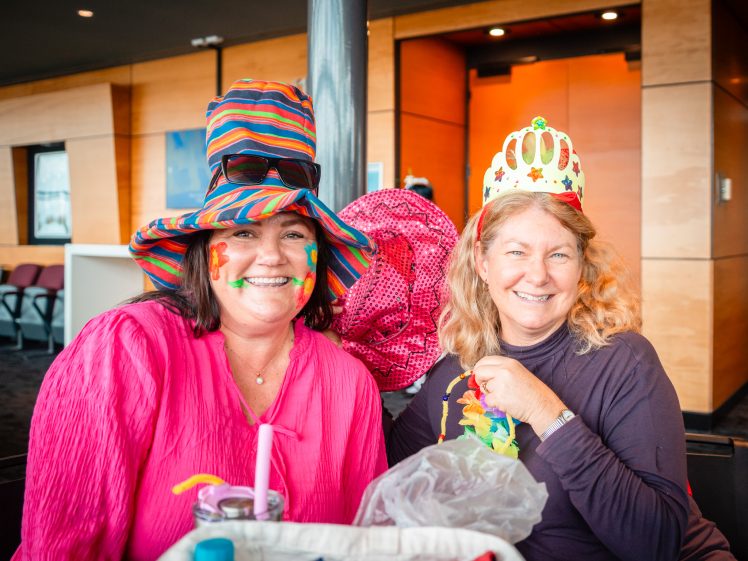 Image of Two women smiling indoors, wearing colourful hats and face paint. One wears a striped hat and pink top; the other has a paper crown and holds a pink hat, both looking cheerful and festive.