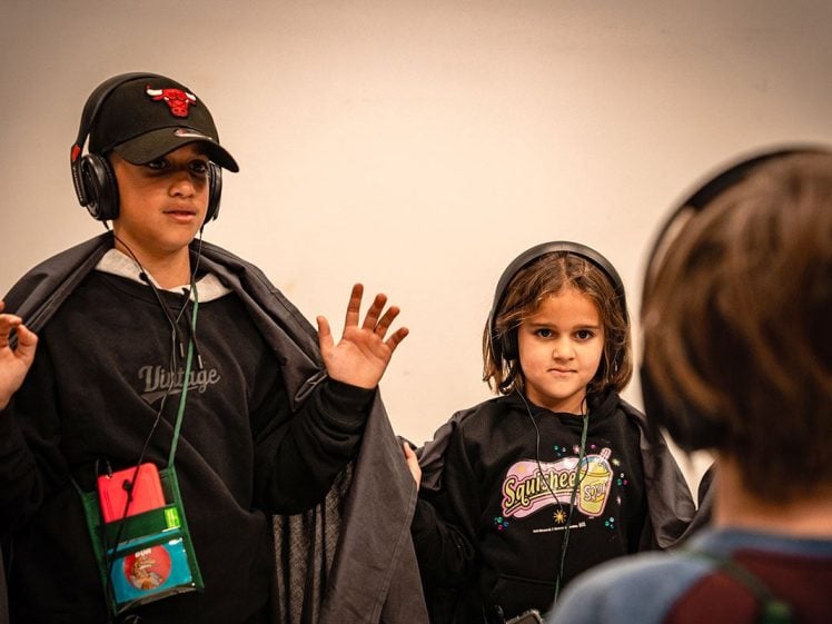 Image of Two children wearing headphones stand together; one raises their hands while wearing a cap and draped in a cloak, and the other stands beside them. Another child with headphones is visible from behind in the foreground.