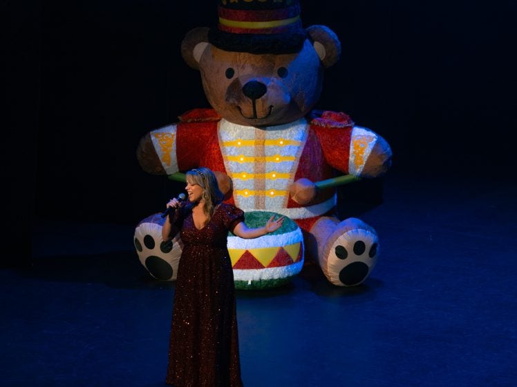 Image of A woman in a sparkling red gown sings on stage, holding a microphone, with a large, colourful inflatable teddy bear drummer in the background under spotlight.