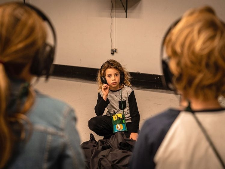 Image of A child sits cross-legged on the floor, listening and holding an earpiece, whilst two other children with headphones face them in the foreground. The setting appears to be an indoor room or studio.