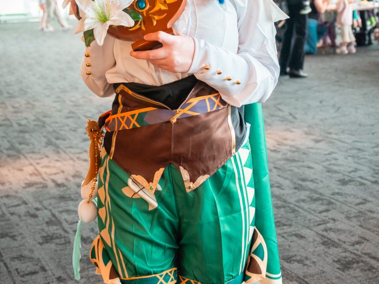 Image of A person in a colourful, detailed cosplay outfit with a white ruffled shirt, green shorts, white tights, and a feathered hat poses indoors, smiling and holding a decorative lyre adorned with flowers.