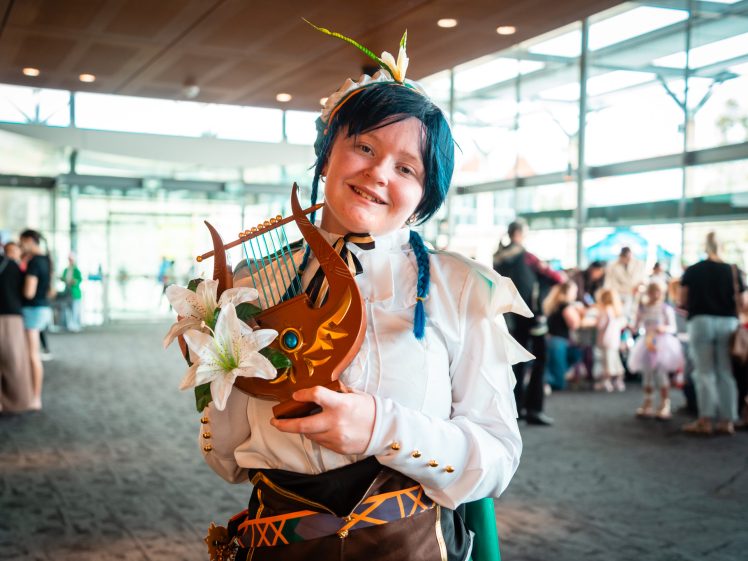 Image of A person in fancy dress, wearing a white frilled shirt, blue wig, and a flower crown, smiles whilst holding a decorative lyre with white lilies. They stand in a large, sunlit indoor space with people in the background.