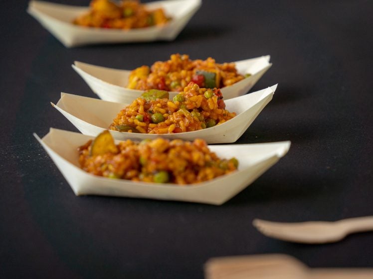 Image of Three small wooden trays filled with rice and vegetables are arranged in a line on a dark surface, with two wooden forks beside them in the foreground.
