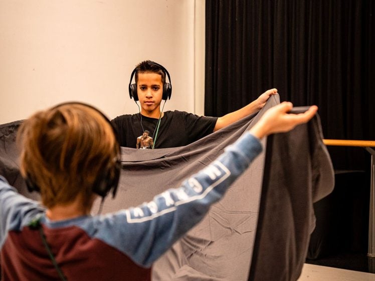 Image of A child in the foreground holds up a large grey cloth, facing an adult wearing headphones in a room with white walls and a black curtain. Both individuals appear to be engaged in an interactive activity.
