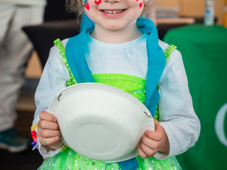 Image of A young child wearing a crown and a green dress with silver stars smiles at the camera. Their face is painted with red hearts and colourful designs, and they hold a white paper plate.