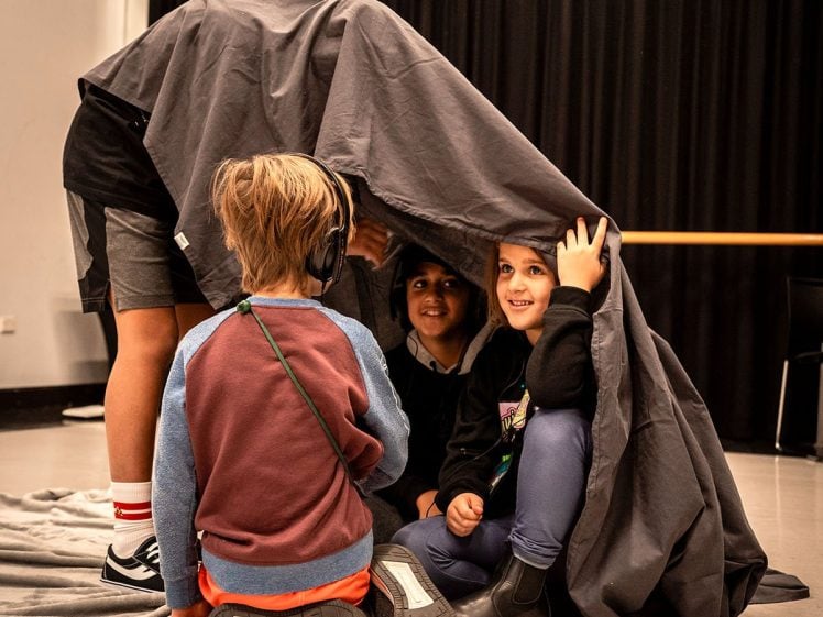 Image of Three children sit on the floor inside a makeshift tent made from a grey blanket, smiling and looking out. An adult stands outside, holding the blanket up. The scene is set in a room with black curtains and light-coloured walls.