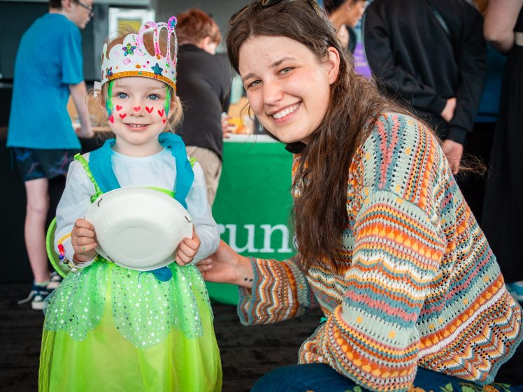 Image of A smiling young girl in a green princess dress, crown, and face paint stands next to a smiling woman in a colourful jumper. The girl holds a paper plate, and people are visible in the background at an indoor event.
