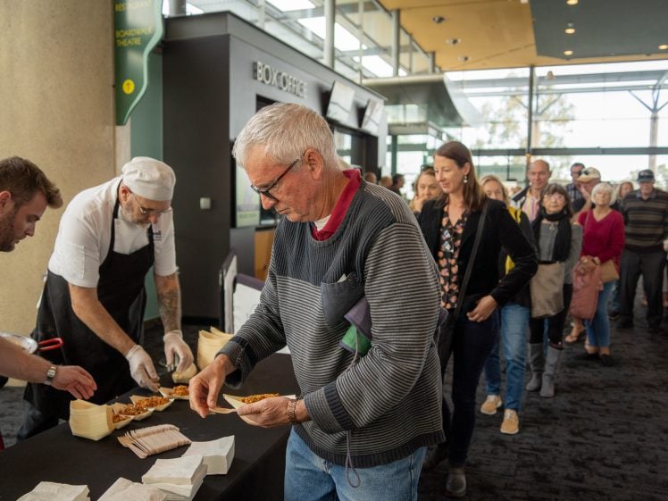 Image of A group of people queue at an indoor event as two staff in chef uniforms serve food from behind a table. A man in glasses at the front takes a slice of pizza, while others wait behind him.