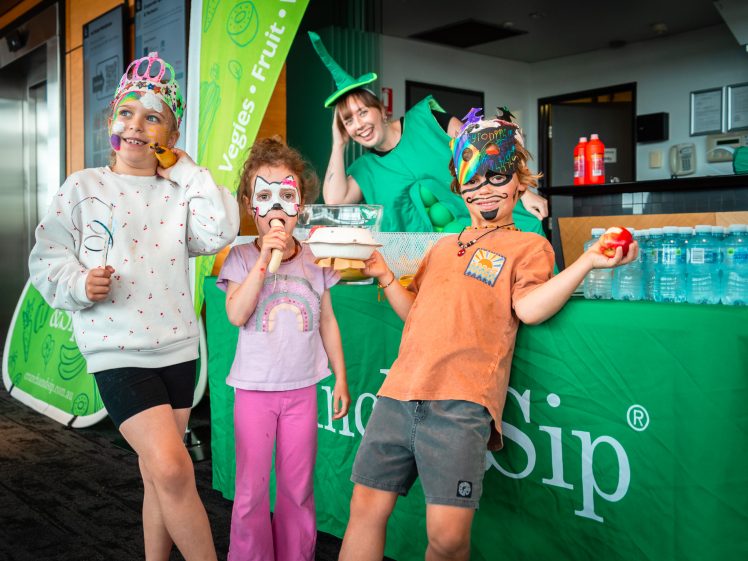 Image of Three children in colourful masks and costumes pose happily in front of a green table with drinks, joined by an adult in a vegetable costume, promoting healthy snacks and drinks indoors.