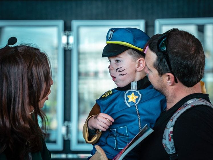 Image of A young child dressed in a police officer fancy dress with face paint is being held by an adult, whilst another adult speaks to them. The child looks towards the adult, and they are indoors with glass doors in the background.