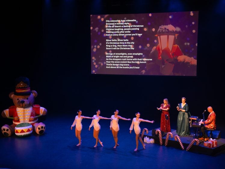 Image of Five dancers in reindeer costumes perform on stage with a large festive bear, two women singing near a keyboard, and Christmas decorations. Song lyrics are projected on a screen in the background.