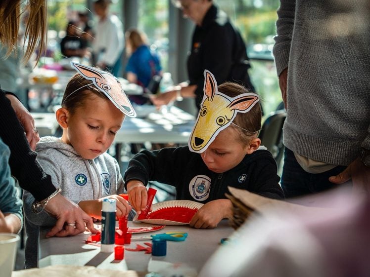 Image of Two young children wearing animal face masks concentrate on a craft project at a busy table, surrounded by adults and art supplies in a bright, indoor setting.