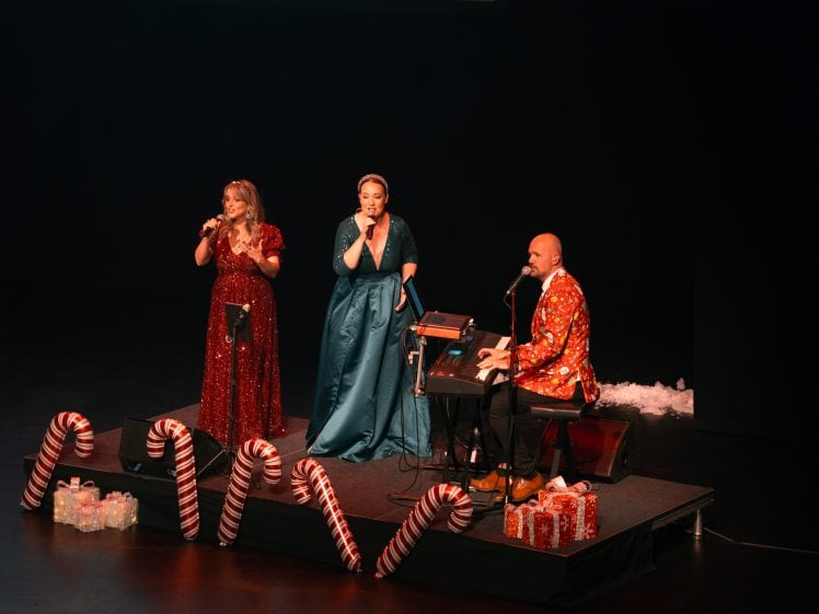 Image of Three performers onstage: two women in elegant dresses singing into microphones and a man in a festive suit playing keyboard. The stage is decorated with large candy canes, artificial snow, and wrapped presents, creating a festive atmosphere.