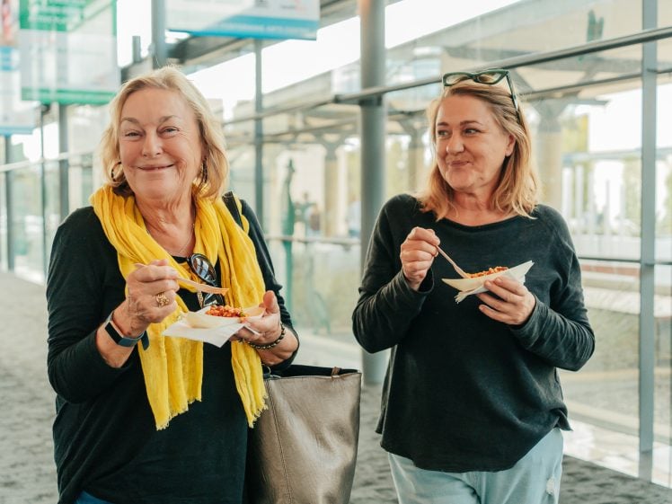 Image of Two women standing indoors, smiling and eating food from small trays with forks. One wears a yellow scarf and holds a large bag; the other has sunglasses on her head. Bright, natural light streams through windows behind them.