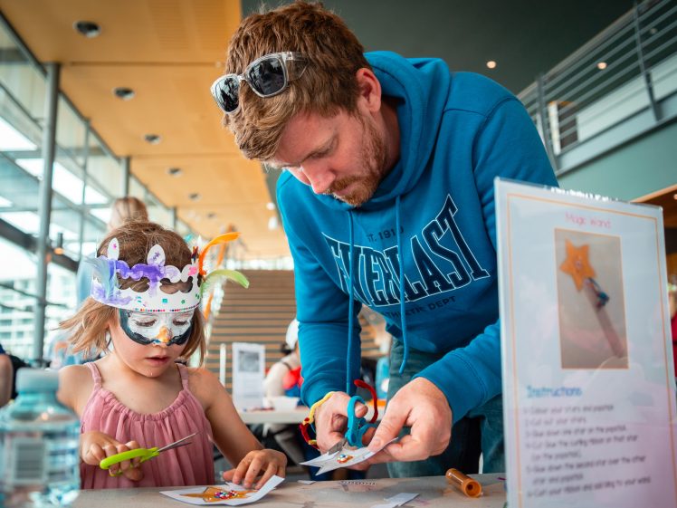 Image of A bearded man helps a young girl wearing a colourful handmade mask with arts and crafts at a table. They are both cutting paper, and the girl is focused on her task. Other people and bright indoor lighting are visible in the background.