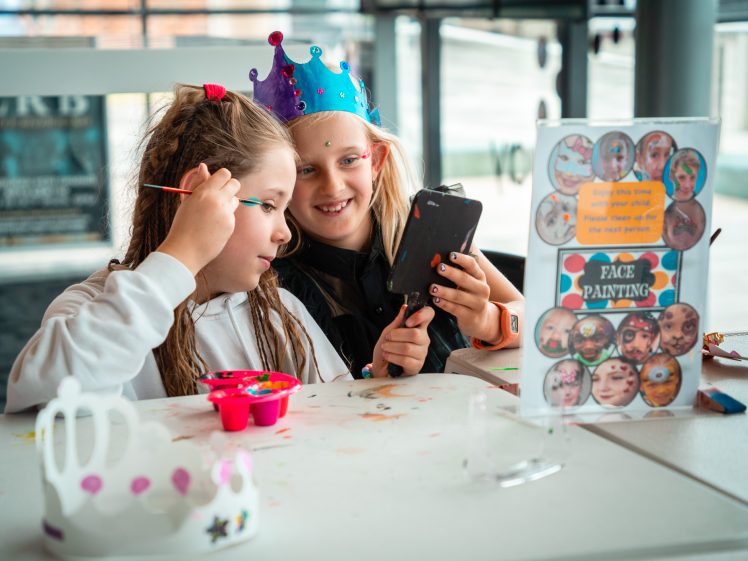 Image of Two young girls wearing colourful crowns smile as one paints the other's face. They look into a handheld mirror. A sign on the table reads Face Painting, and painting supplies are scattered about.