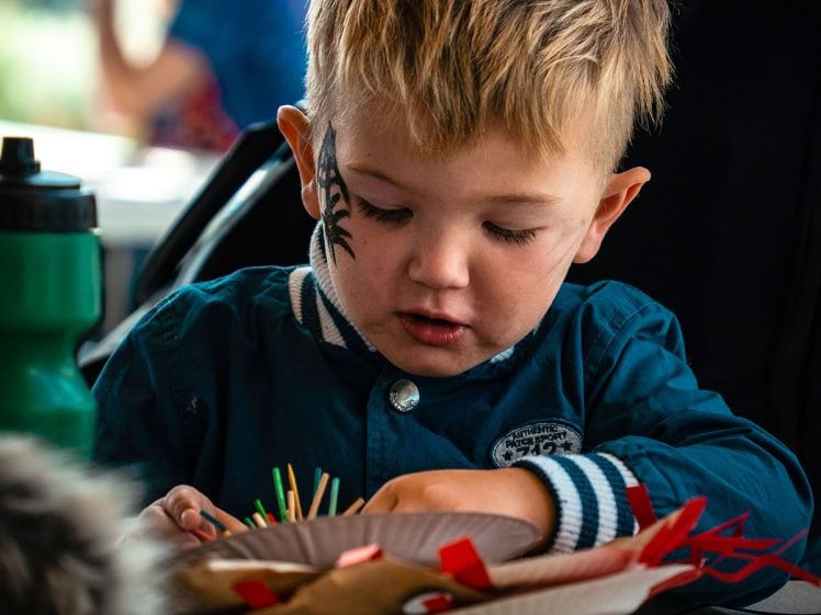 Image of A young boy with face paint concentrates on a craft project, picking sticks from a paper plate. He wears a blue jacket and sits at a table, with a green water bottle nearby.