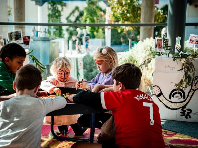 Image of Four young children sit together around a low table, playing with toys. One child has face paint, and another wears a headband. Sunlight streams through large windows, and decorated boxes and plants are visible in the background.