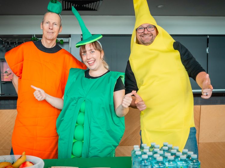 Image of Three adults in carrot, pea pod, and banana costumes stand smiling behind a table with water bottles, bananas, and brochures, indoors at a cheerful event.