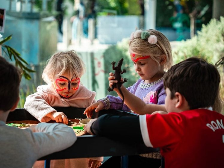 Image of Four young children play at a table covered in sand and toys. Two girls with face paint are focused on the activity, one holding a toy tree. A boy in a red shirt sits nearby. Bright daylight and greenery surround them.