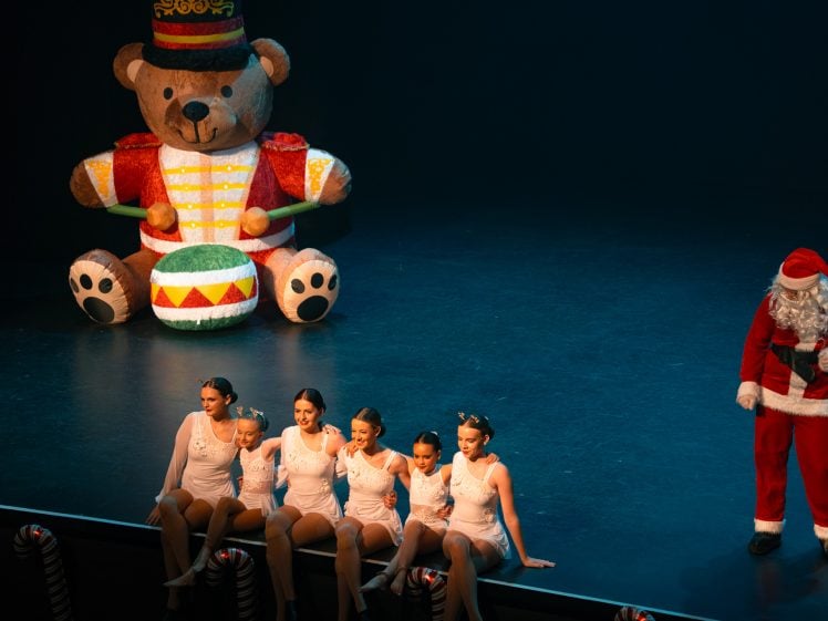 Image of Five dancers in white costumes sit on stage, smiling, next to a person dressed as Father Christmas. An inflatable toy soldier bear holding a drum is in the background. The stage is decorated with candy cane props.