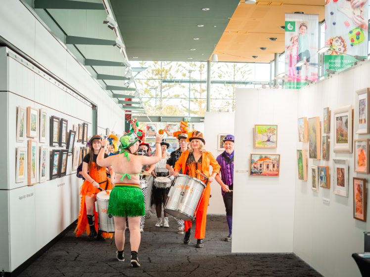 Image of A group of musicians in colourful, festive costumes plays drums in an art gallery, following a person in a green hula skirt and leafy headpiece. Paintings are displayed on the gallery walls. Bright natural light fills the space.