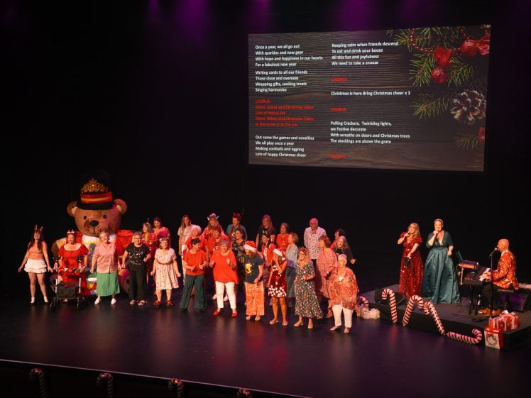 Image of A group of performers in festive costumes sing on stage with holiday decorations. A large teddy bear and candy canes are visible, with Christmas song lyrics projected on a screen behind them.
