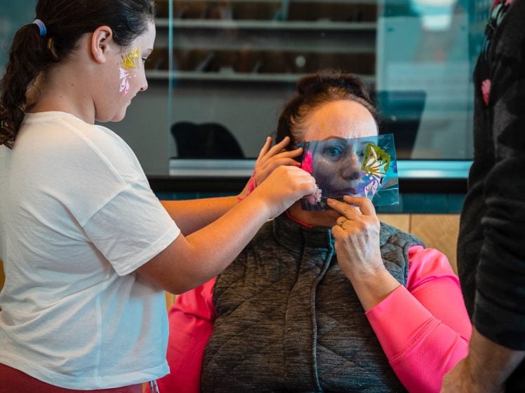 Image of A child applies a colourful face sticker to a seated woman’s face while an adult stands nearby. Art supplies and a mirror are on the table in front of them, and the scene appears to be in a creative workshop space.