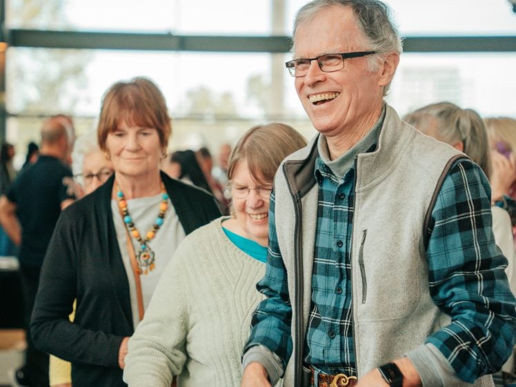 Image of A group of older adults stands indoors, smiling and talking. One man in the foreground wears glasses, a grey waistcoat, and a checked shirt, appearing cheerful. Other people are gathered in the brightly lit background.