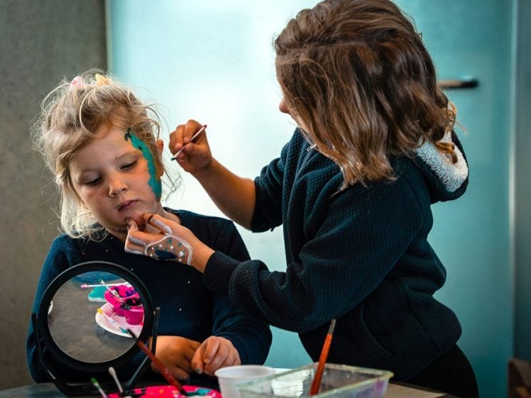 Image of Two young children sit at a table; one is painting the other's face with blue and green paint while the other sits still. A mirror and art supplies are on the table in front of them.