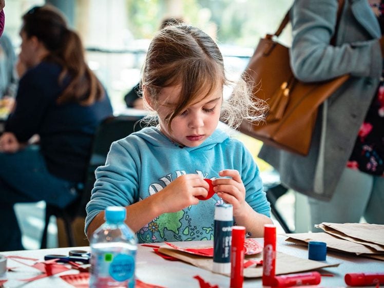 Image of A young girl in a blue hoodie sits at a table focused on crafting with red paper and glue sticks, surrounded by supplies and other people in a bright, busy room.