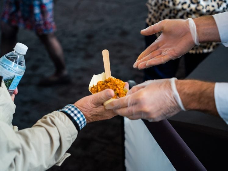 Image of A person wearing gloves hands a small container of food with a wooden spoon to another person holding a water bottle. The receiver is wearing a beige jacket and checked shirt. Only their hands and arms are visible.