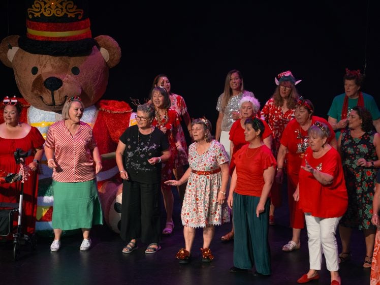 Image of A group of women in festive clothing sing on stage in front of a large teddy bear decoration, celebrating together under bright stage lights.