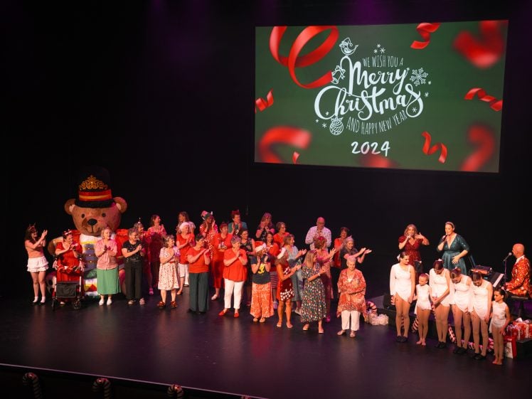 Image of A large group of people in festive attire stand on a stage, some singing, with a person in a bear costume. A screen behind them reads We Wish You a Merry Christmas and a Happy New Year 2024 with festive decorations.