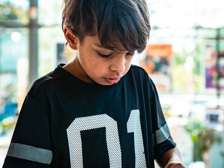 Image of Young boy with brown hair wearing a black shirt with the number 01 focuses intently whilst using his hands to work on a craft project at a table with paper plates and cocktail sticks in a brightly lit room.