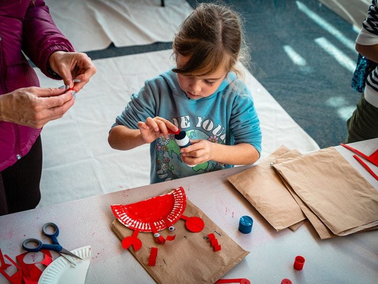 Image of A young girl at a table glues red paper shapes onto a painted paper plate, making a craft. An adult assists nearby. Brown paper bags, scissors, and other crafting materials are on the table.