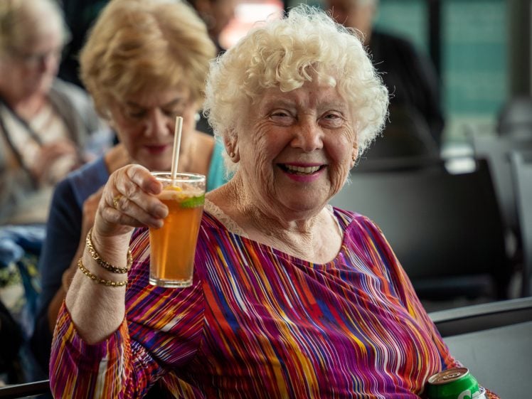Image of Smiling elderly woman with curly white hair holds up a drink in a clear glass with a straw and mint leaves. She wears a colourful striped top and sits indoors, with another woman in the background.