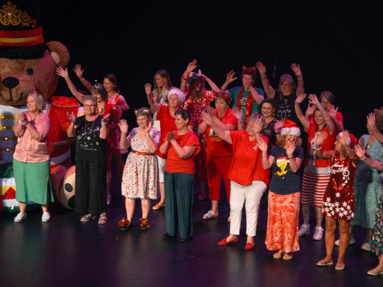 Image of A group of people dressed in colourful, festive outfits stand on stage, waving and smiling. A large costumed bear is in the background, and the atmosphere is joyful and celebratory.