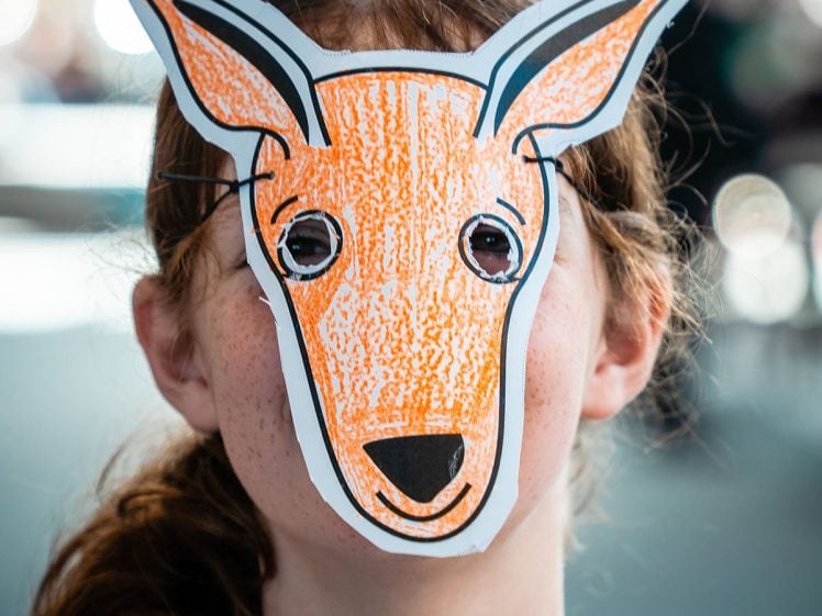 Image of A child wearing a handmade orange and white deer mask, with eye holes cut out, stands outside. The mask covers the child's face completely, and their reddish hair and tie-dye shirt are visible.