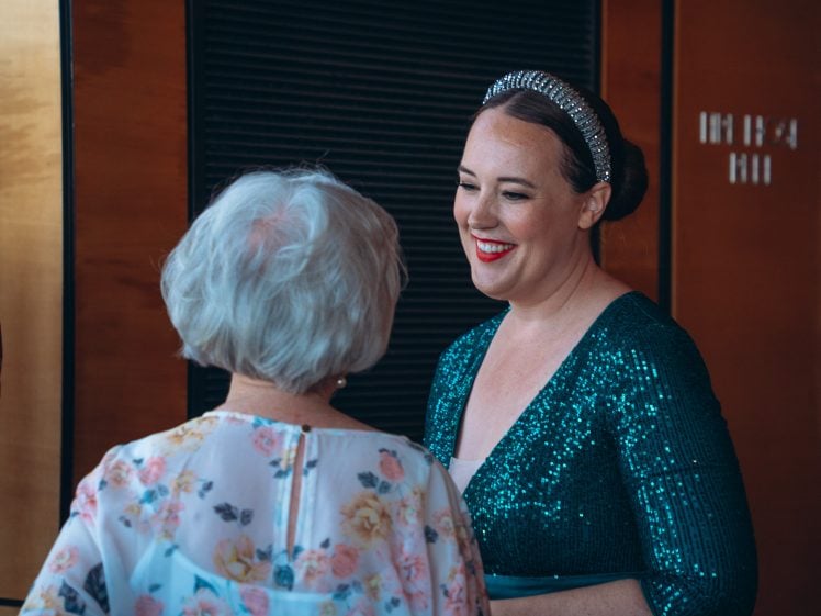 Image of A woman in a sparkly green dress and jewelled headband smiles and talks with an older woman with short white hair, who is wearing a floral blouse. They are indoors with warm lighting.