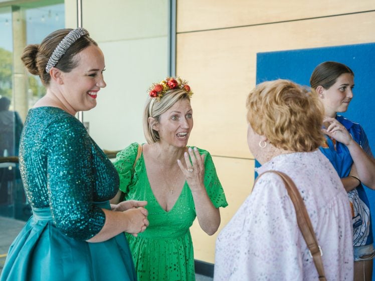Image of Three women are talking and smiling at an indoor event. One wears a sparkly green dress, another has a flower crown and a green dress, and the third has a pink top. A fourth woman stands in the background wearing blue.
