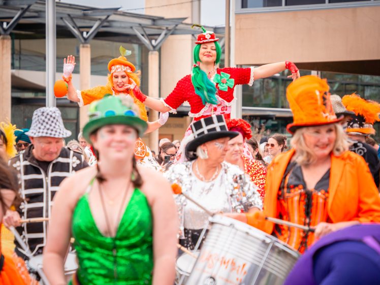 Image of A lively parade features people in colourful costumes, including women in green, orange, and red outfits, with hats and festive accessories, smiling and playing drums in an outdoor urban setting.