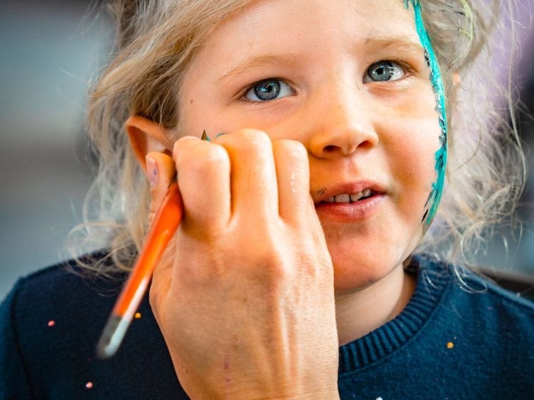 Image of A young child with blonde hair and blue eyes sits still while someone paints green designs on their face using a brush. The child wears a navy blue shirt with small colourful dots.