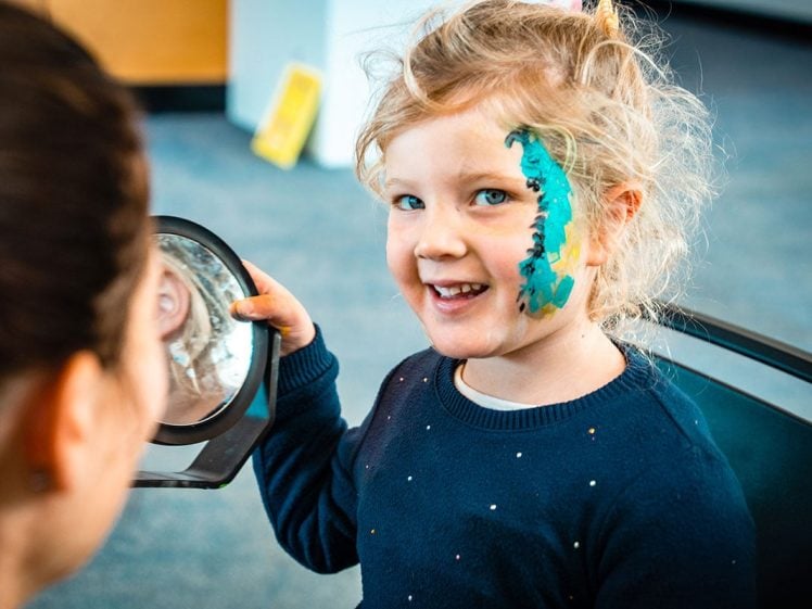 Image of A young child with curly blonde hair smiles whilst looking into a handheld mirror, showing off a face paint design of a green dragon on their cheek. An adult is partially visible in the foreground.