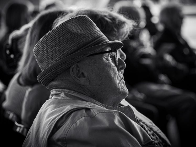 Image of An elderly man wearing a hat and glasses sits in profile among a group of people, appearing thoughtful. The photo is in black and white, with others blurred in the background.