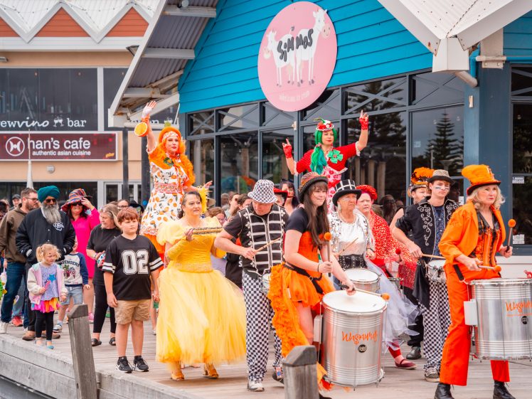 Image of A lively parade of people in colourful costumes, including clowns, musicians, and children, walks along a promenade outside shops and restaurants, creating a festive and cheerful atmosphere.