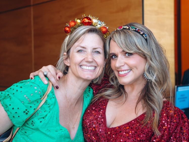 Image of Two women smile together indoors. One wears a green dress and a festive headband with ornaments; the other wears a red sequinned dress and a jewelled headband. They appear happy and close, posing for the photo.